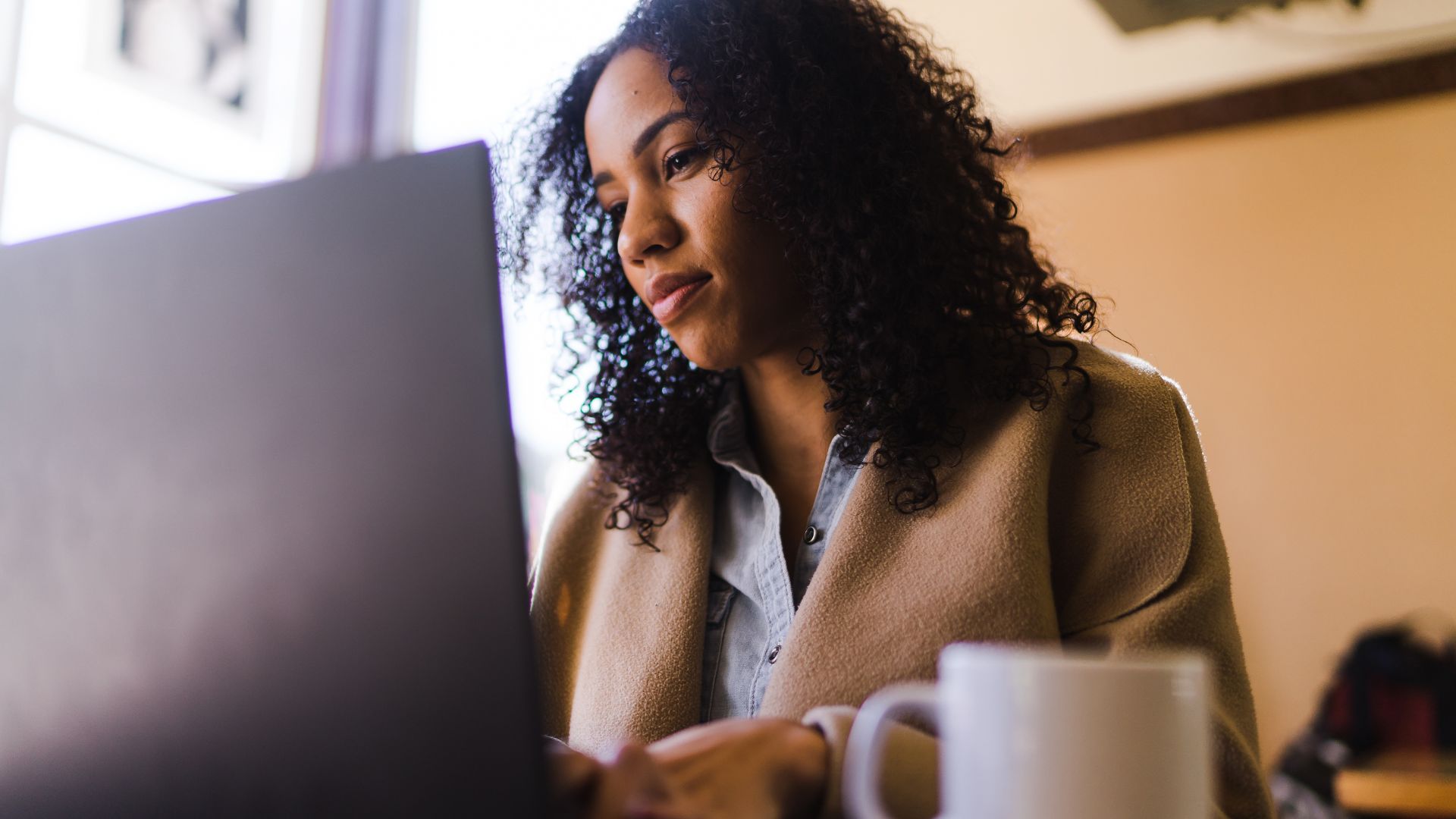 Woman with curly hair working on a laptop at a desk with a coffee mug nearby.